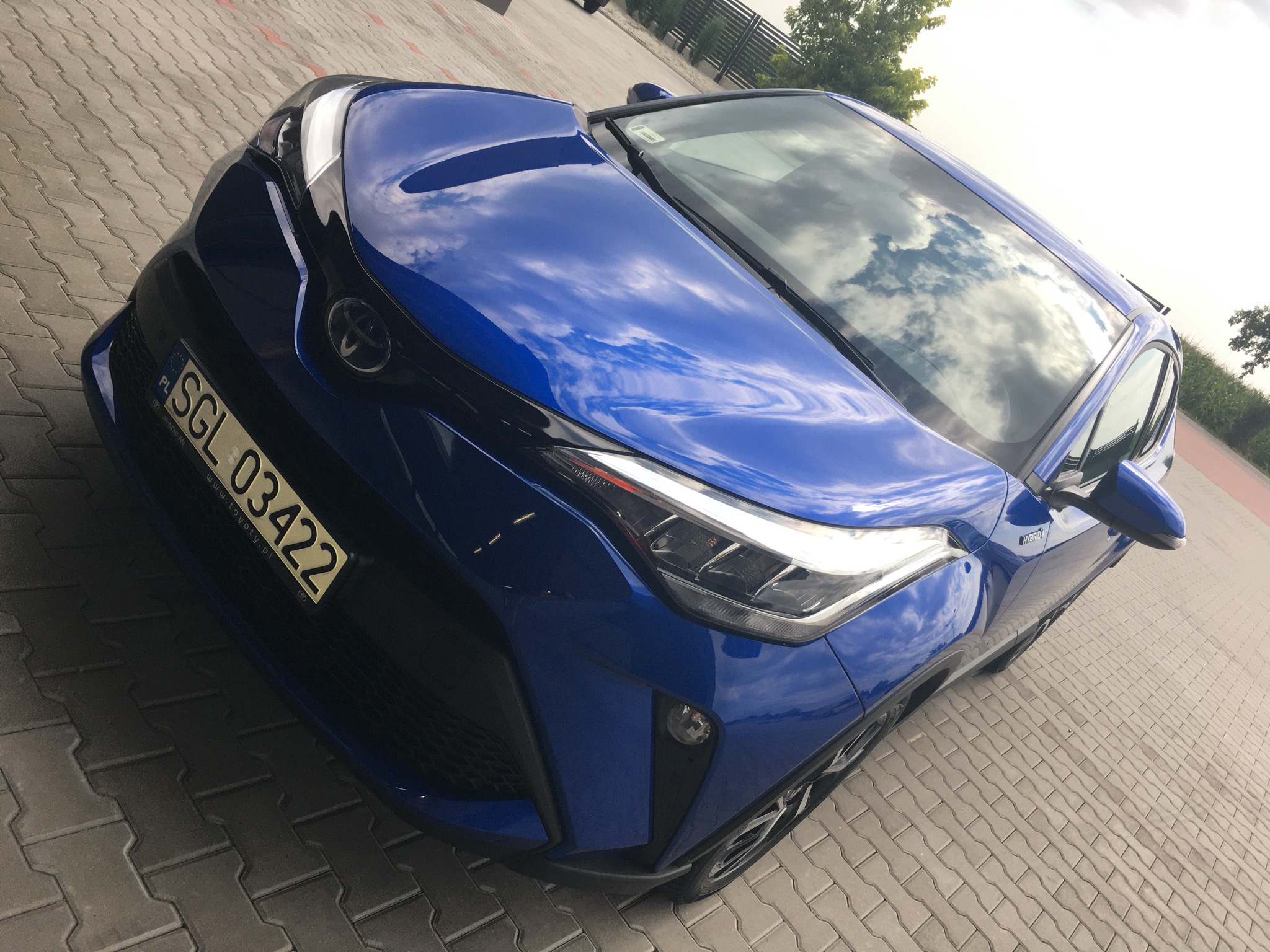 Błyszczący, niebieski samochód Toyota C-HR z widocznym odbiciem chmur na lakierze, zaparkowany na kostce brukowej. Tablica rejestracyjna SGL 03422.