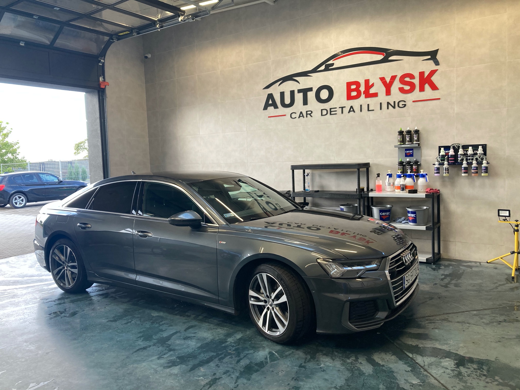 Szary sedan Audi po usłudze detailingu, widoczny w jasnym garażu z logo firmy Auto Błysk Car Detailing na ścianie i akcesoriami do pielęgnacji samochodów w tle.