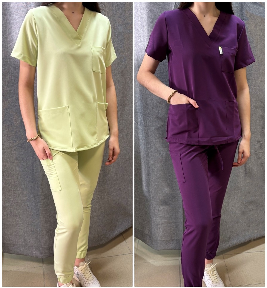 Dwa komplety odzieży medycznej: pistacjowy i fioletowy. Bluzy z krótkim rękawem i spodnie ze ściągaczami. Modelki z rękami w kieszeniach, na szarym tle.
