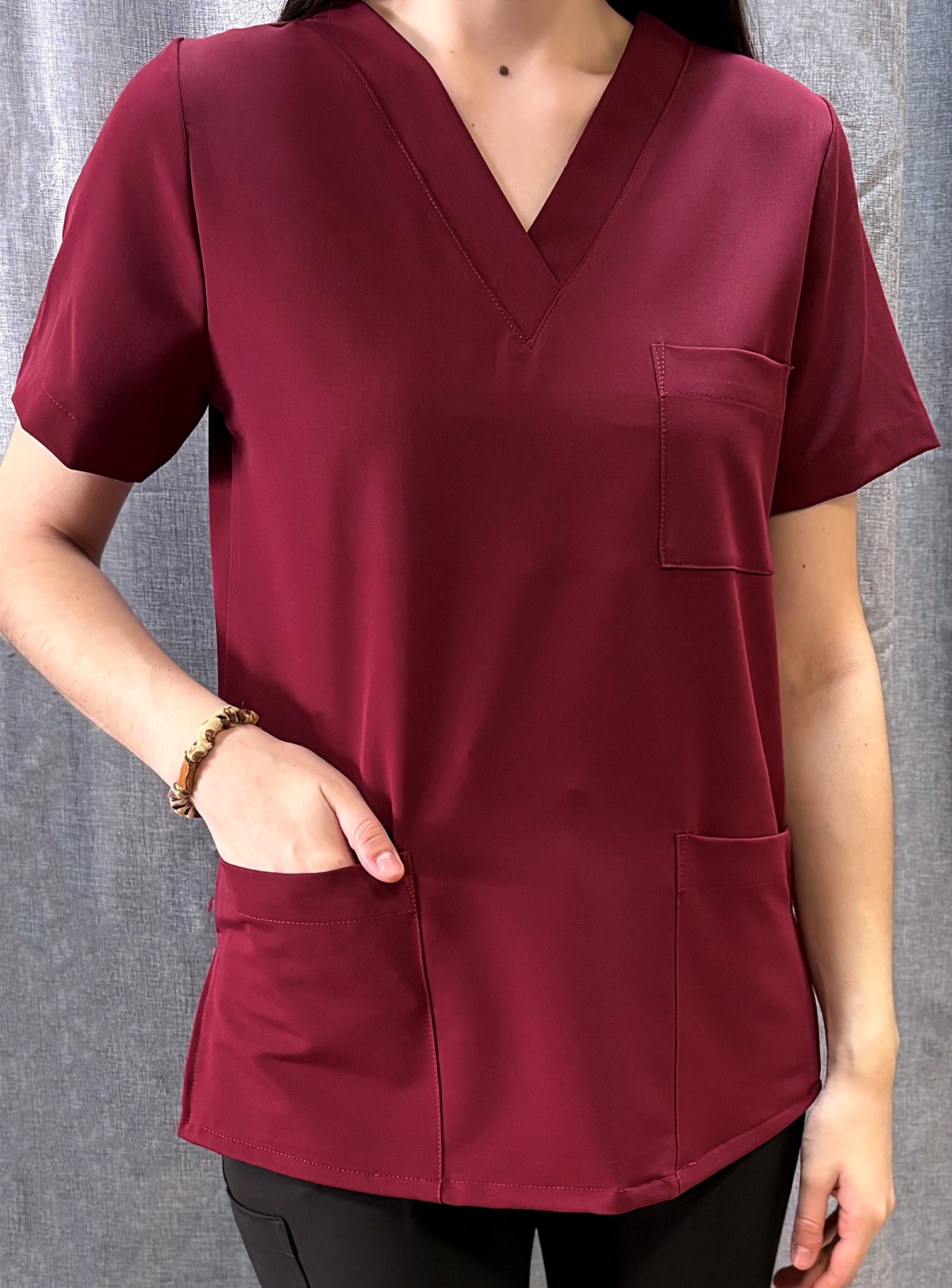 Burgundowa bluza medyczna z kieszeniami, prezentowana na osobie z bransoletką. Widoczne detale szycia i materiału na tle szarej zasłony.