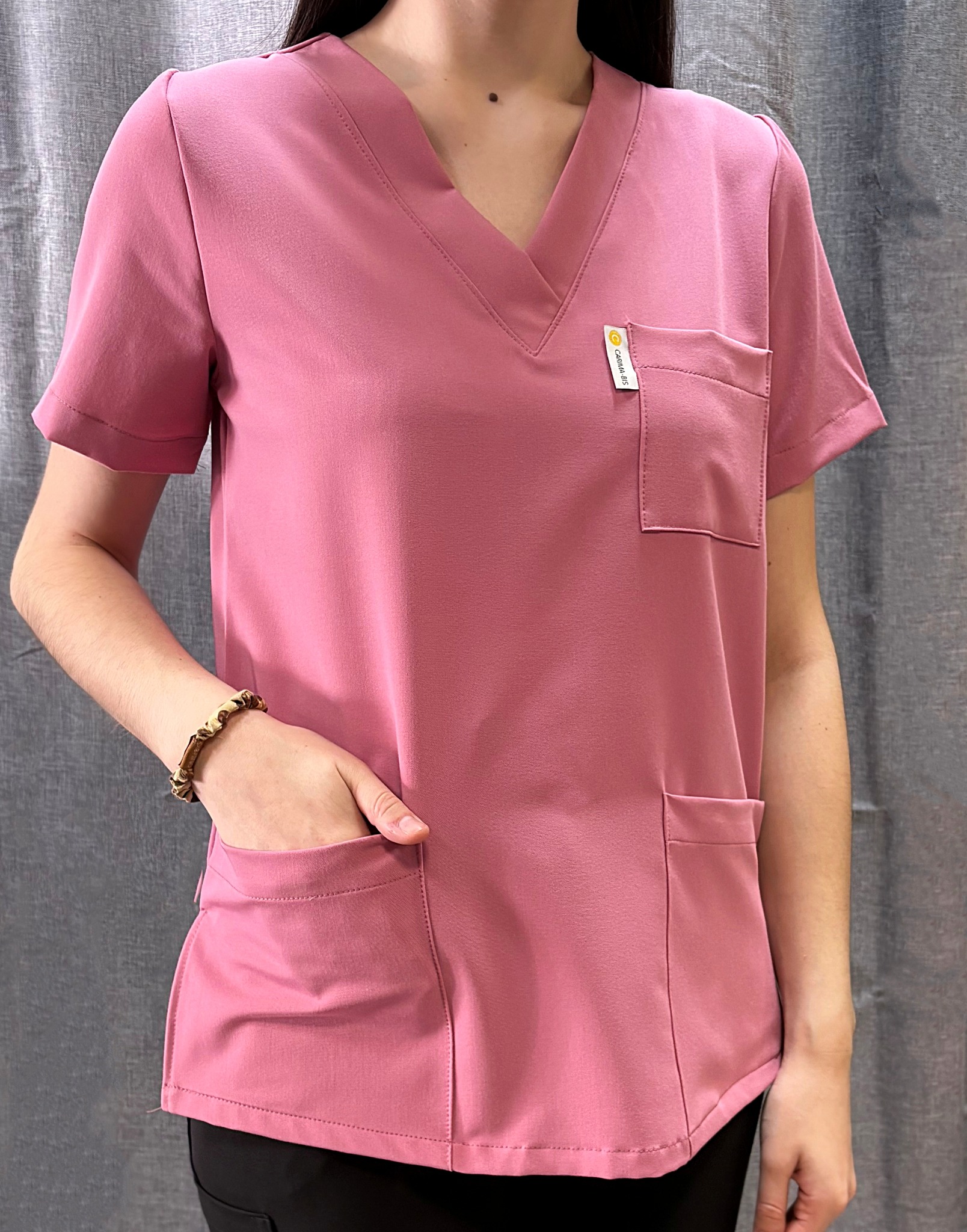 Różowa bluza medyczna z krótkim rękawem, kieszeniami i logo na tle szarej zasłony. Osoba ma rękę w kieszeni.