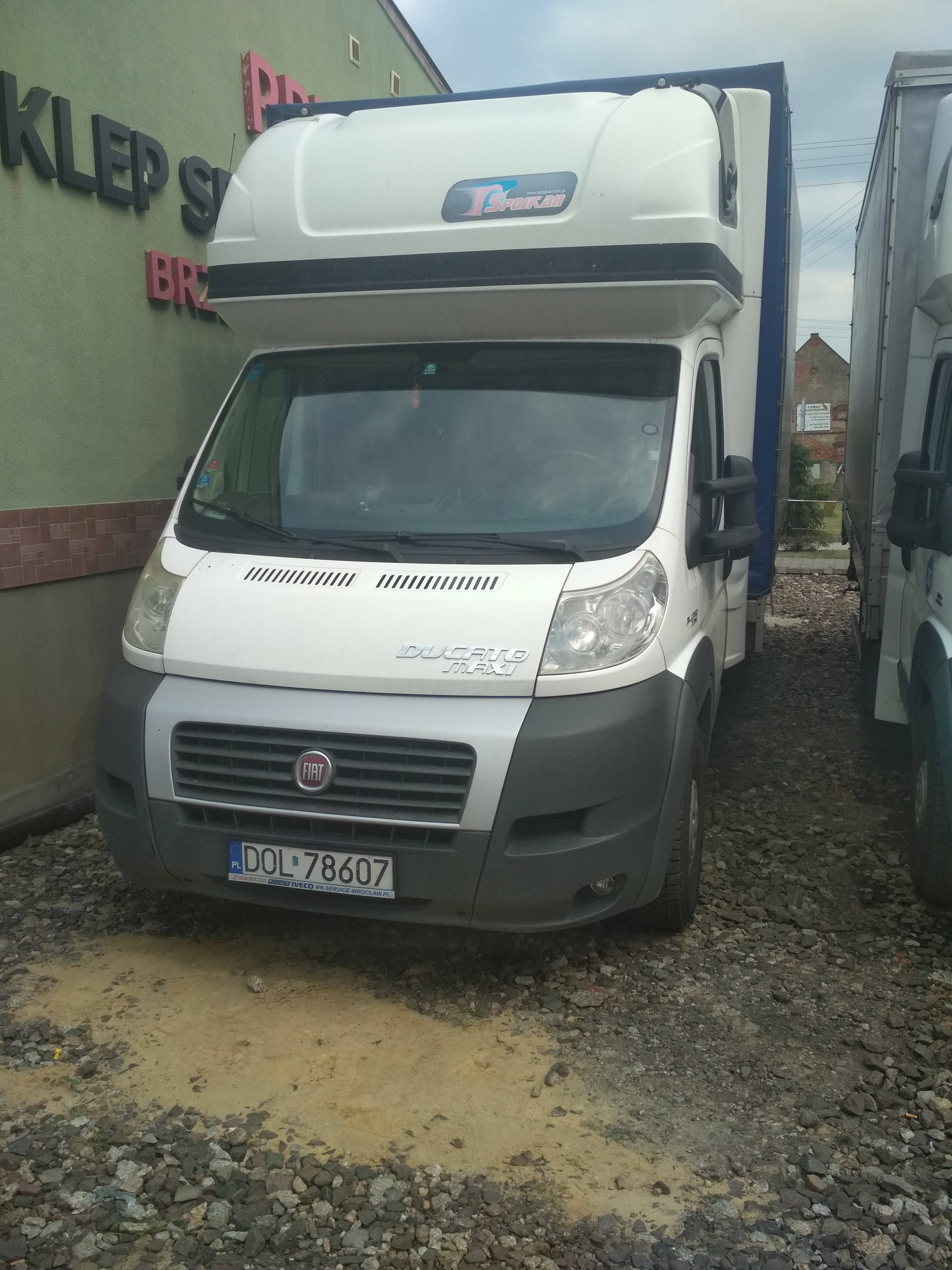 Biały bus dostawczy Fiat Ducato Maxi z zabudową kontenerową, zaparkowany na żwirowym placu, widoczny znak firmowy 'Spojkar' na kontenerze.