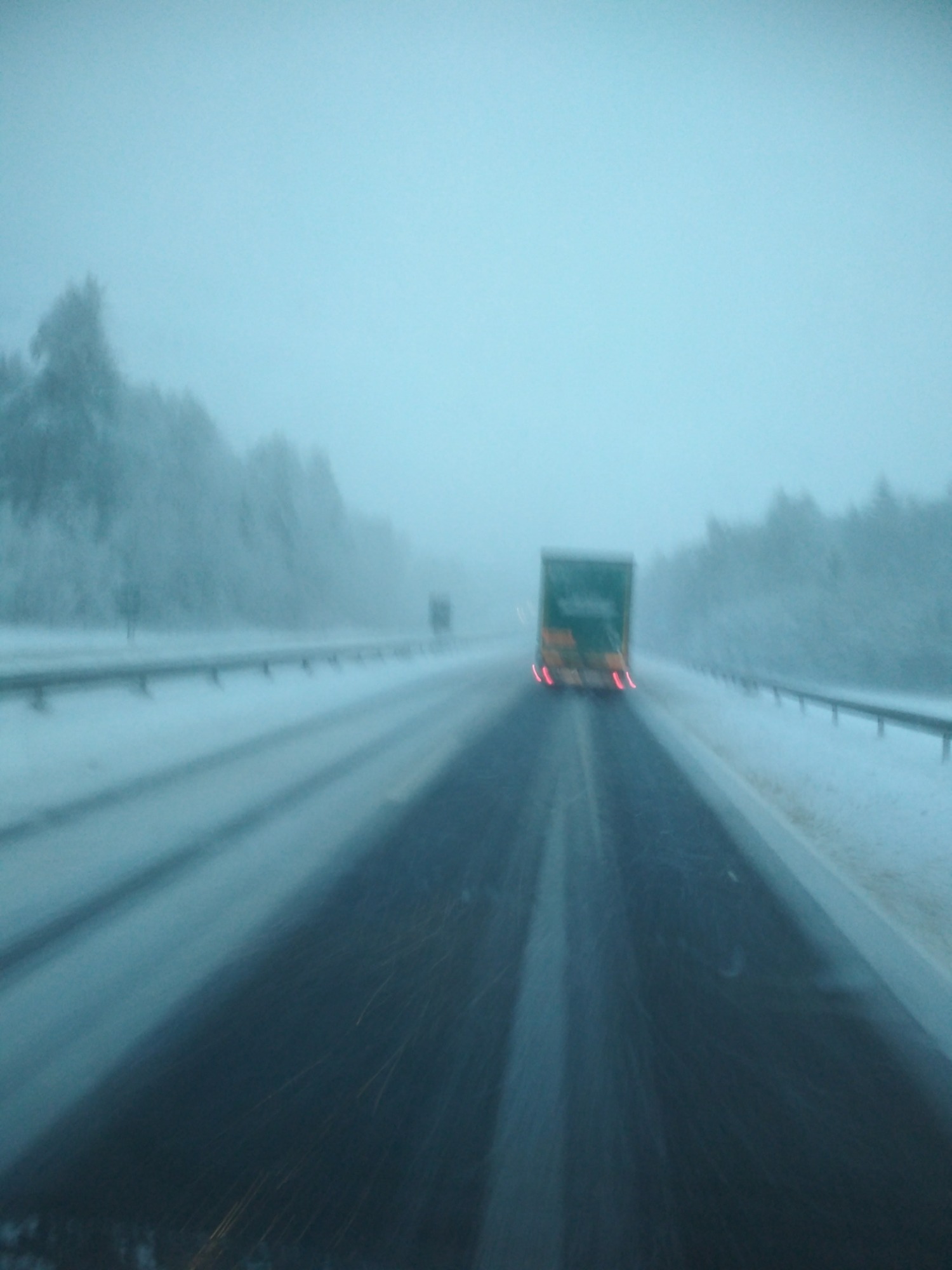 Ciężarówka z włączonymi światłami hamowania jedzie zasypaną śniegiem autostradą w warunkach ograniczonej widoczności.