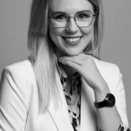 Portret uśmiechniętej blondynki w okularach, białym żakiecie i wzorzystej bluzce, opierającej brodę na dłoni z widocznym zegarkiem na bransolecie.