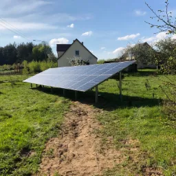 Panel słoneczny zamontowany na metalowej konstrukcji na zielonej łące, w tle dom jednorodzinny i błękitne niebo z chmurami.