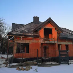 Surowy budynek jednorodzinny w trakcie budowy, z częściowo położonym ciemnoszarym dachem, widoczne rusztowania i zasypany śniegiem teren.