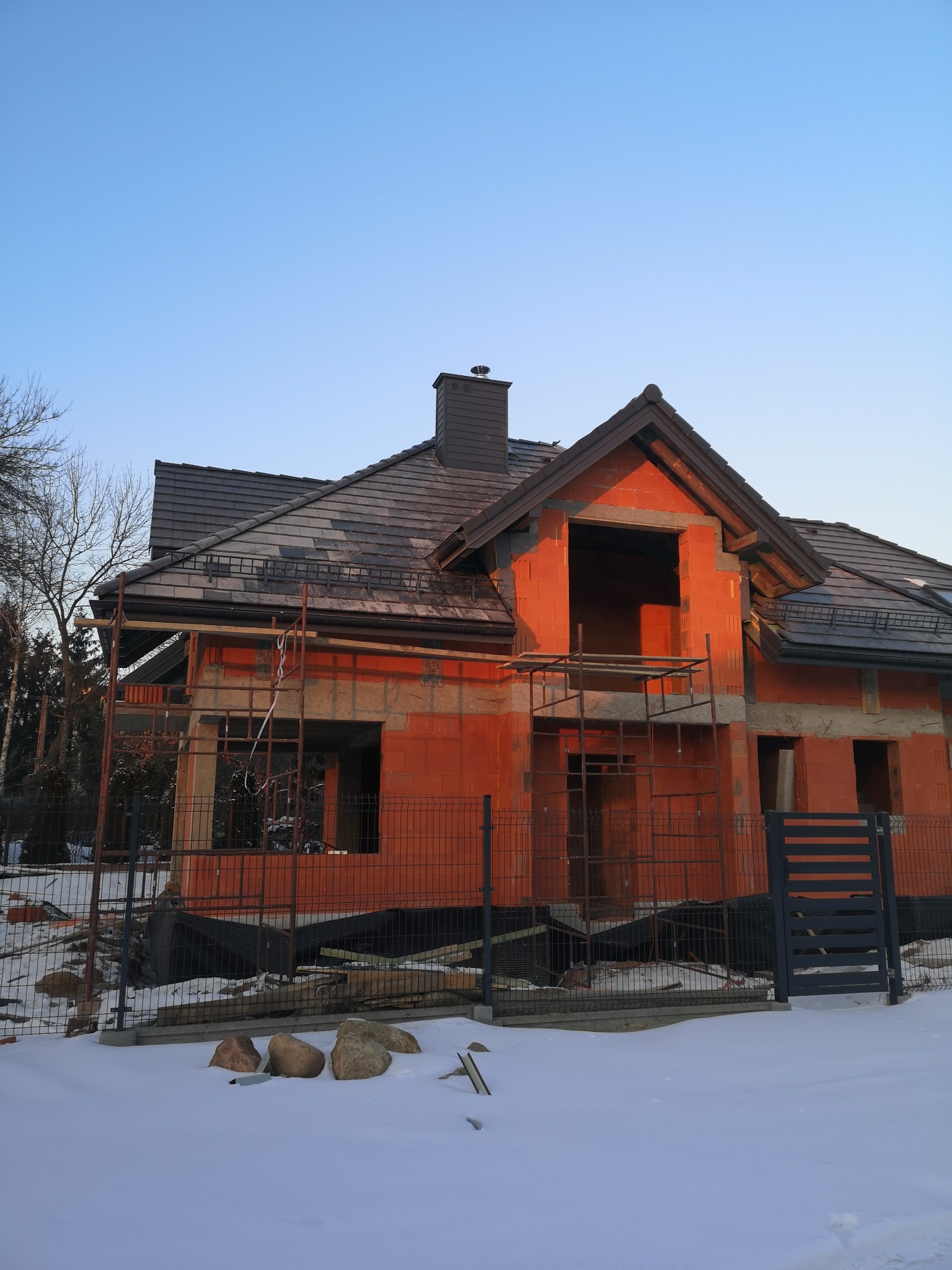 Surowy budynek jednorodzinny w trakcie budowy, z częściowo położonym ciemnoszarym dachem, widoczne rusztowania i zasypany śniegiem teren.