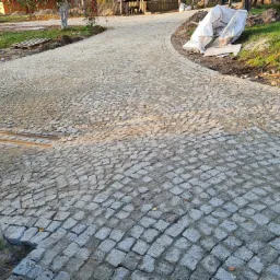 Świeżo ułożona, zakrzywiona ścieżka z kostki granitowej w ogrodzie, widoczne przygotowane podłoże z elementami szyn oraz worki z materiałem budowlanym w tle.