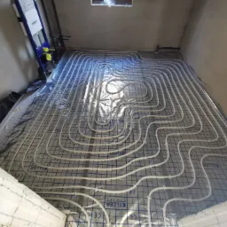 Białe rurki ogrzewania podłogowego ułożone na folii z nadrukiem Keller System, widok z góry na surową podłogę w trakcie instalacji, z fragmentem stelaża podtynkowego Geberit w tle.