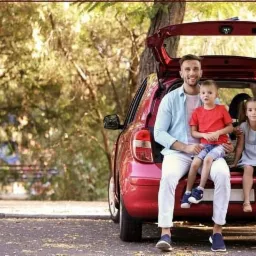 Uśmiechnięta rodzina z dwójką dzieci siedzi na bagażniku czerwonego samochodu podczas pikniku w słoneczny dzień, w tle drzewa, w lewym górnym rogu logo firmy ubezpieczeniowej.