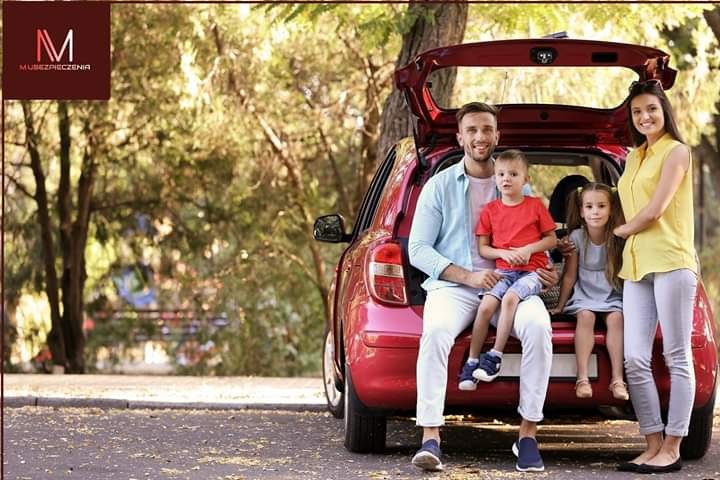 Uśmiechnięta rodzina z dwójką dzieci siedzi na bagażniku czerwonego samochodu podczas pikniku w słoneczny dzień, w tle drzewa, w lewym górnym rogu logo firmy ubezpieczeniowej.