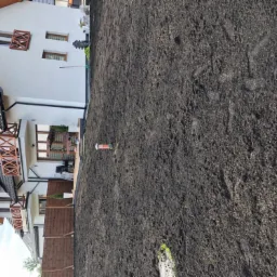 Przygotowany teren pod trawnik w nowym budownictwie, widoczna świeżo nawieziona ziemia, ogrodzenie i fragmenty budynków w tle. Ślady stóp na ziemi.