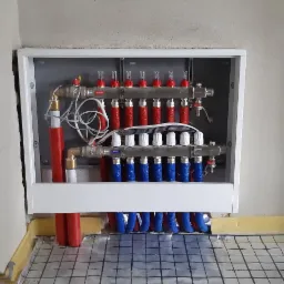 Rozdzielacz ogrzewania podłogowego w białej obudowie, widoczne rury w izolacji czerwonej i niebieskiej, mata grzewcza na podłodze, surowy stan ścian.