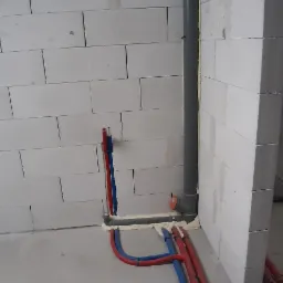 Wewnętrzna instalacja wodna i kanalizacyjna w surowym stanie: rury czerwone i niebieskie prowadzone po podłodze i ścianie z betonu komórkowego, zabezpieczone pianką montażową.