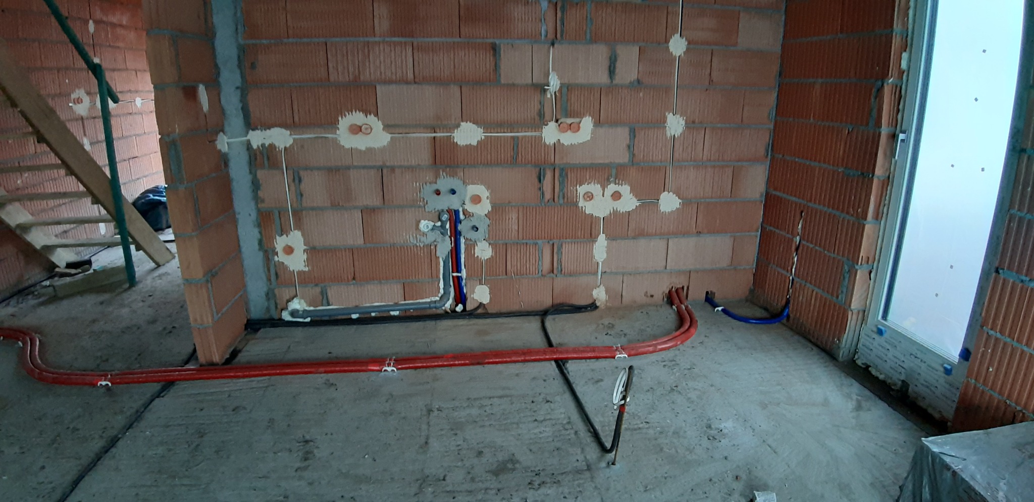 Instalacja wodna i elektryczna w surowym wnętrzu budynku z czerwonej cegły, widoczne rury w podłodze i wyprowadzenia ze ściany, przygotowane do dalszych prac wykończeniowych.