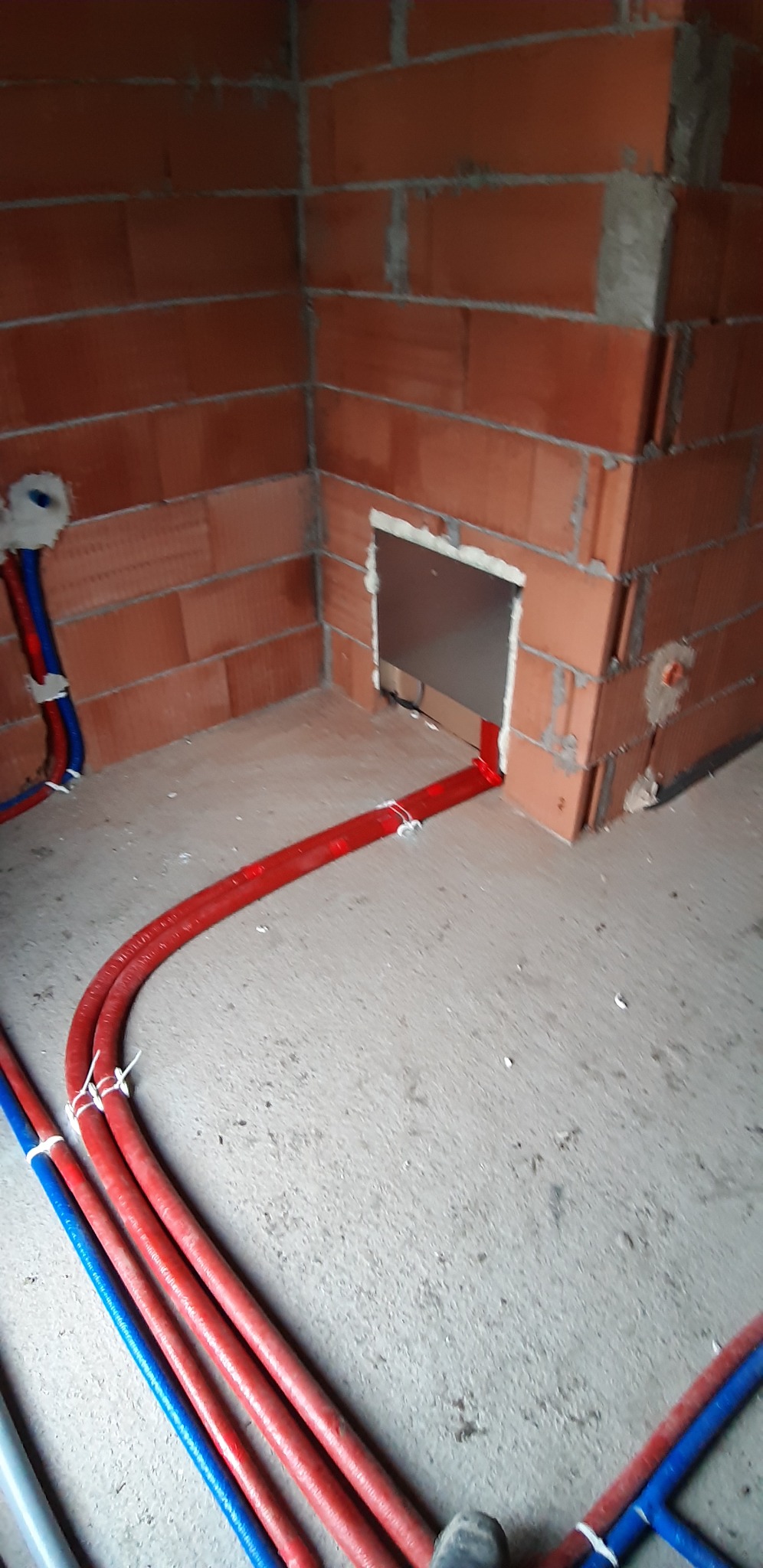 Wewnętrzny widok instalacji w surowym stanie: czerwone i niebieskie rury grzewcze prowadzone po podłodze wzdłuż ściany z pustaków ceramicznych, z otworem w ścianie zabezpieczonym pianką montażową.