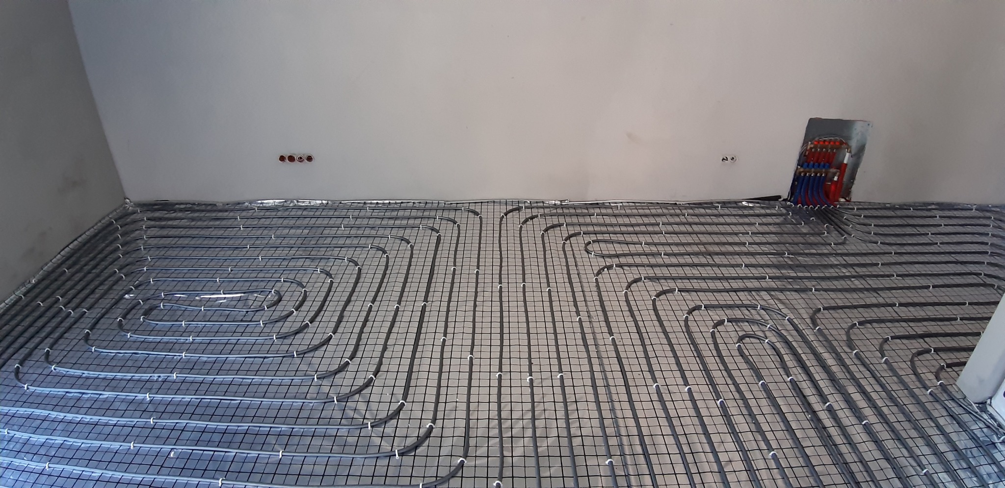 Rozłożone rury ogrzewania podłogowego na srebrnej folii izolacyjnej, widoczny rozdzielacz z rurami doprowadzającymi wodę w kolorach czerwonym i niebieskim, surowy stan wykończenia ścian