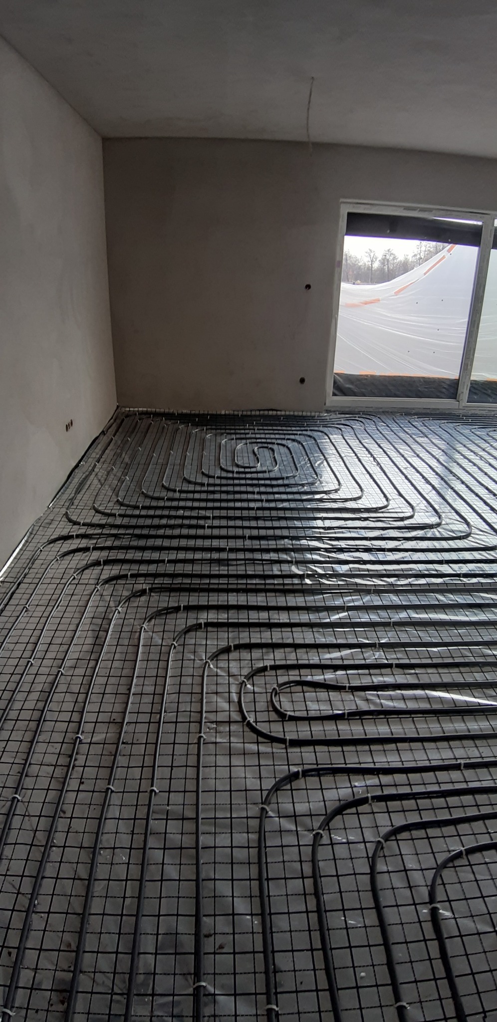 Rozłożona instalacja ogrzewania podłogowego na siatce w surowym wnętrzu, widoczne rury grzewcze układane meandrycznie przed wylewką, okno z widokiem na zewnątrz.
