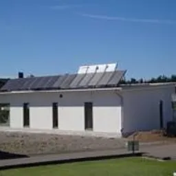 Biały, parterowy dom jednorodzinny z płaskim dachem, na którym zamontowane są panele słoneczne. Budynek otoczony zielonym trawnikiem i drzewami, w tle widoczny czerwony dom. Minimalistyczna...