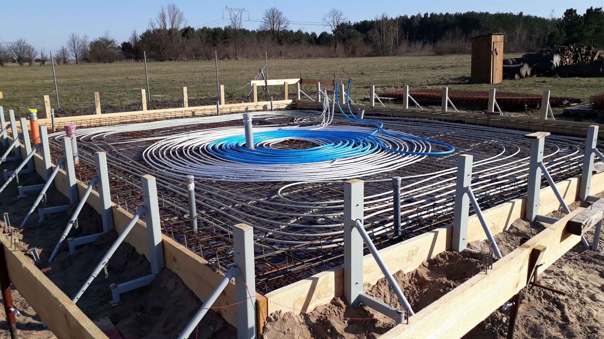 Przygotowanie płyty fundamentowej pod dom zeroenergetyczny: widoczne szalunki, zbrojenie i instalacja ogrzewania podłogowego z rur w kolorze białym i niebieskim, na tle polnego krajobrazu.
