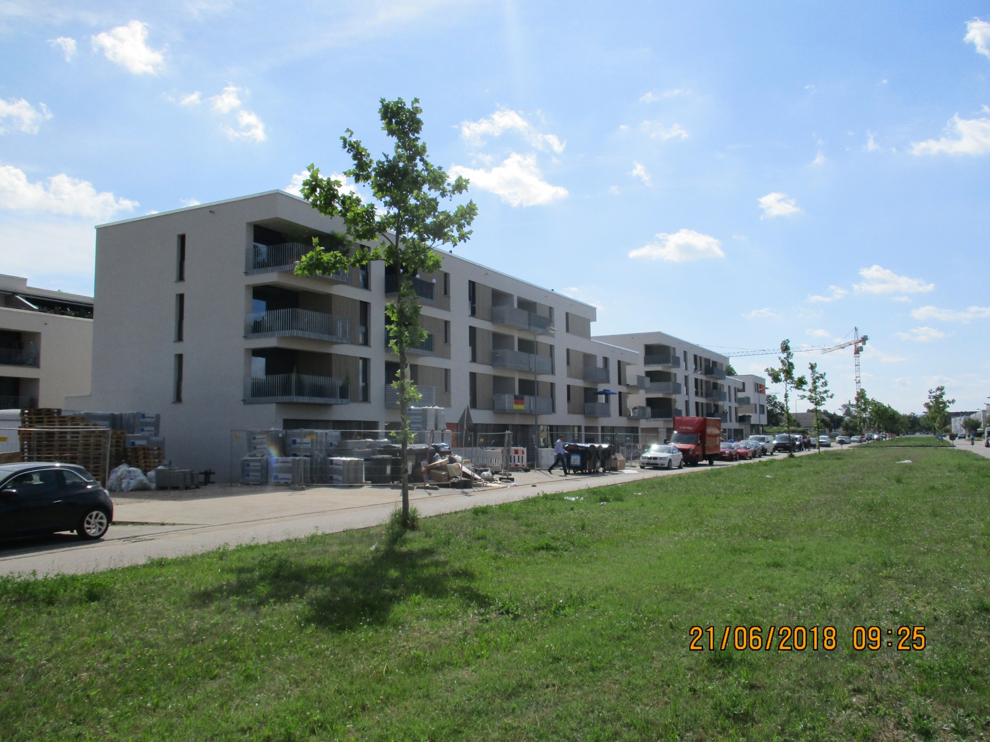 Nowoczesne, trzypiętrowe budynki mieszkalne z balkonami w Karlsruhe, Niemcy, z widocznymi paletami materiałów budowlanych i pracownikami na chodniku przed budynkiem, z dźwigiem w tle.