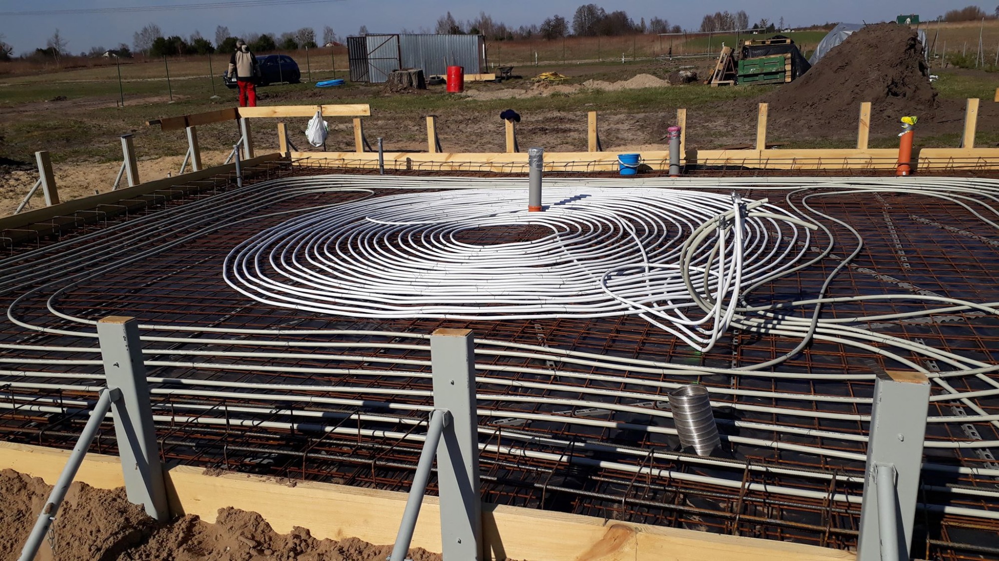 Przygotowanie płyty fundamentowej pod dom zeroenergetyczny: widoczne ułożone rury ogrzewania podłogowego w spiralnym wzorze na siatce zbrojeniowej, wystające pionowe rury instalacyjne, drewniane...