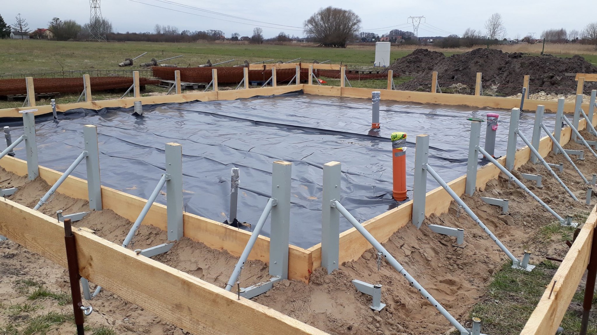 Przygotowanie fundamentów pod budowę domu: drewniany szalunek z metalowymi wspornikami, czarna folia hydroizolacyjna, wystające rury kanalizacyjne, widok na pole i drzewa w tle.