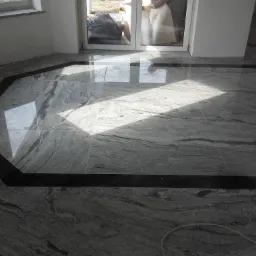 Wykończona podłoga z szarych płyt kamiennych z ciemnym obramowaniem, widoczne okno z parapetem i drzwi balkonowe.