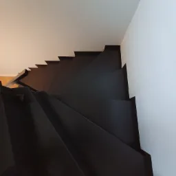 Wysoki kąt widzenia na nowoczesne, ciemne schody z metalową balustradą, kontrastujące z białymi ścianami i sufitem.