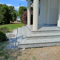 Zewnętrzne schody wejściowe do domu wyłożone jasnoszarym, nakrapianym kamieniem, prowadzące do kolumnady, z widocznymi elementami krajobrazu.