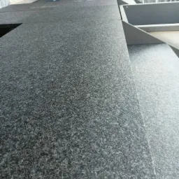 Szorstka, ciemnoszara powierzchnia blatu z widocznym wycięciem, prawdopodobnie pod zlew, w tle elementy konstrukcyjne i fragment zieleni.