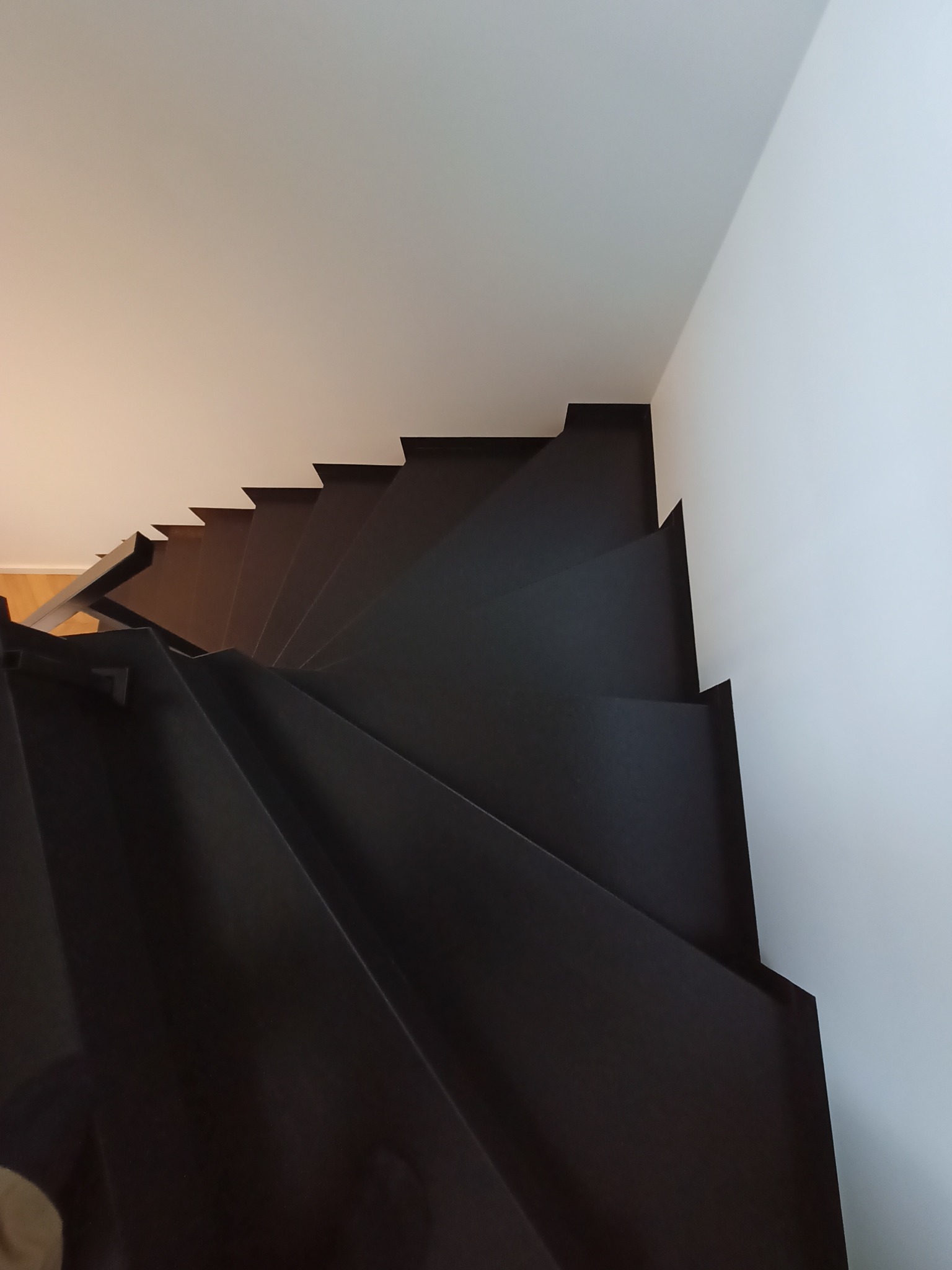 Wysoki kąt widzenia na nowoczesne, ciemne schody z metalową balustradą, kontrastujące z białymi ścianami i sufitem.