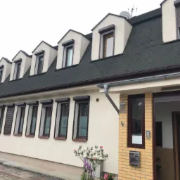 Dom jednorodzinny z zamontowanymi brązowymi roletami zewnętrznymi na oknach, część rolet opuszczona, część podniesiona, widoczny numer domu 68.