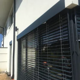 Nowoczesne, grafitowe żaluzje fasadowe zamontowane na elewacji budynku z widocznymi elementami montażowymi i odblaskami otoczenia w lamelach.