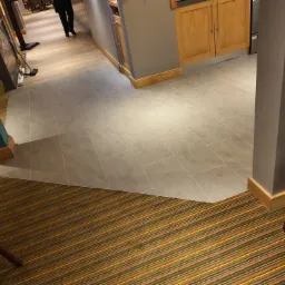 Szara, prostokątna podłoga z płytek ceramicznych w lokalu komercyjnym, przechodząca w brązowo-żółtą wykładzinę dywanową w paski.