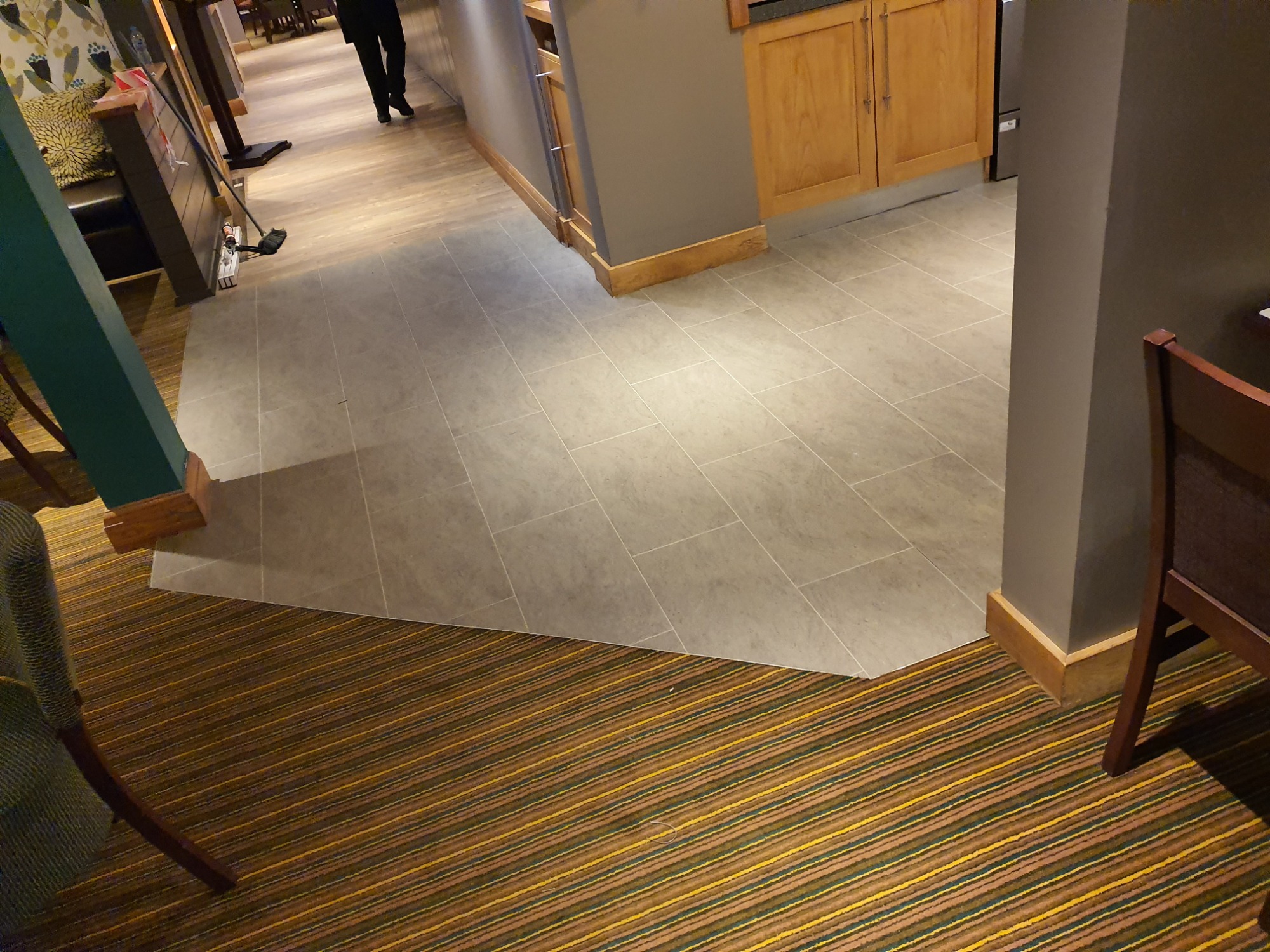 Szara, prostokątna podłoga z płytek ceramicznych w lokalu komercyjnym, przechodząca w brązowo-żółtą wykładzinę dywanową w paski.