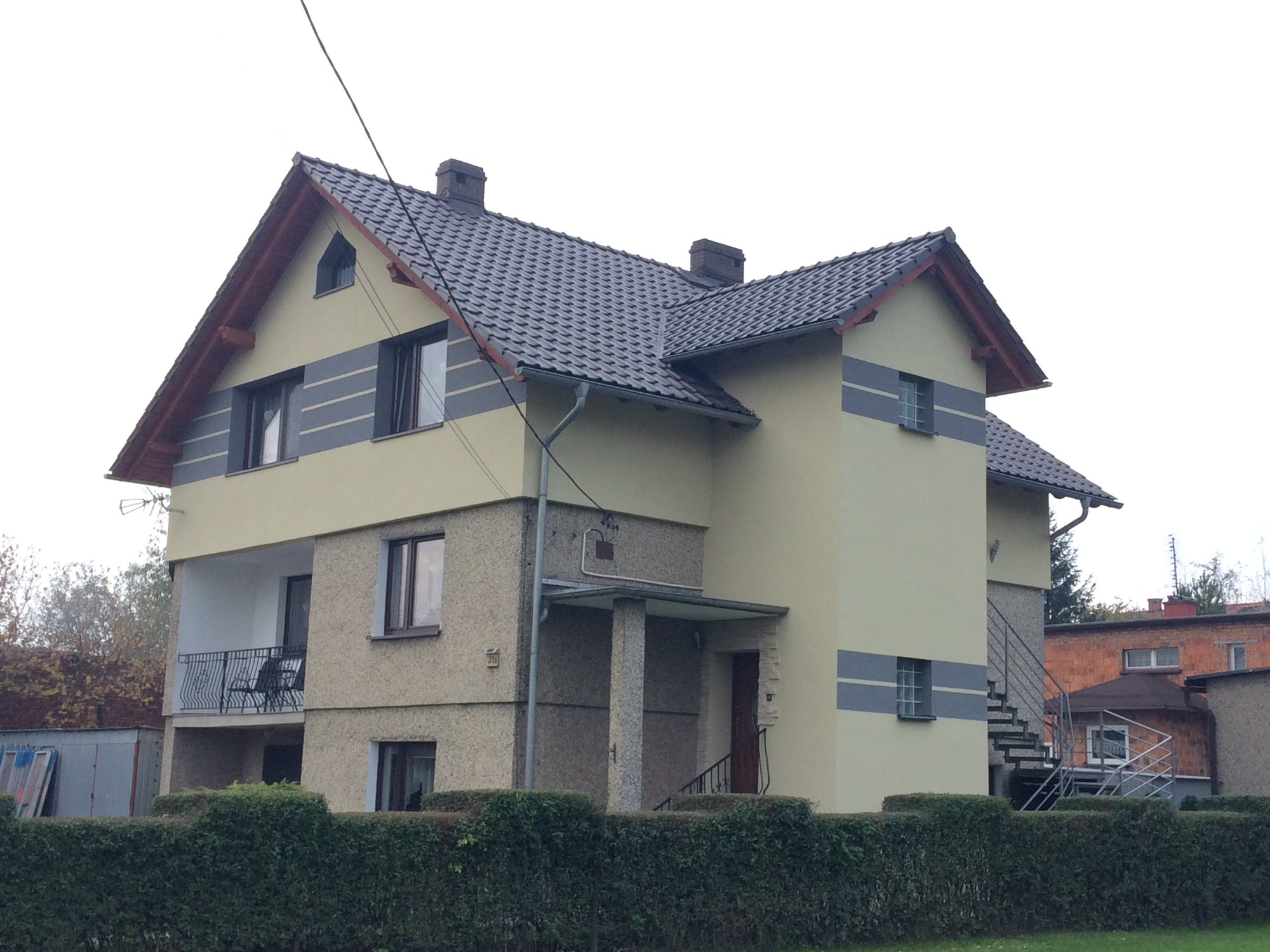 Dwupiętrowy dom z balkonem i zewnętrznymi schodami, elewacja w odcieniach beżu i szarości, dach z ciemnej dachówki.