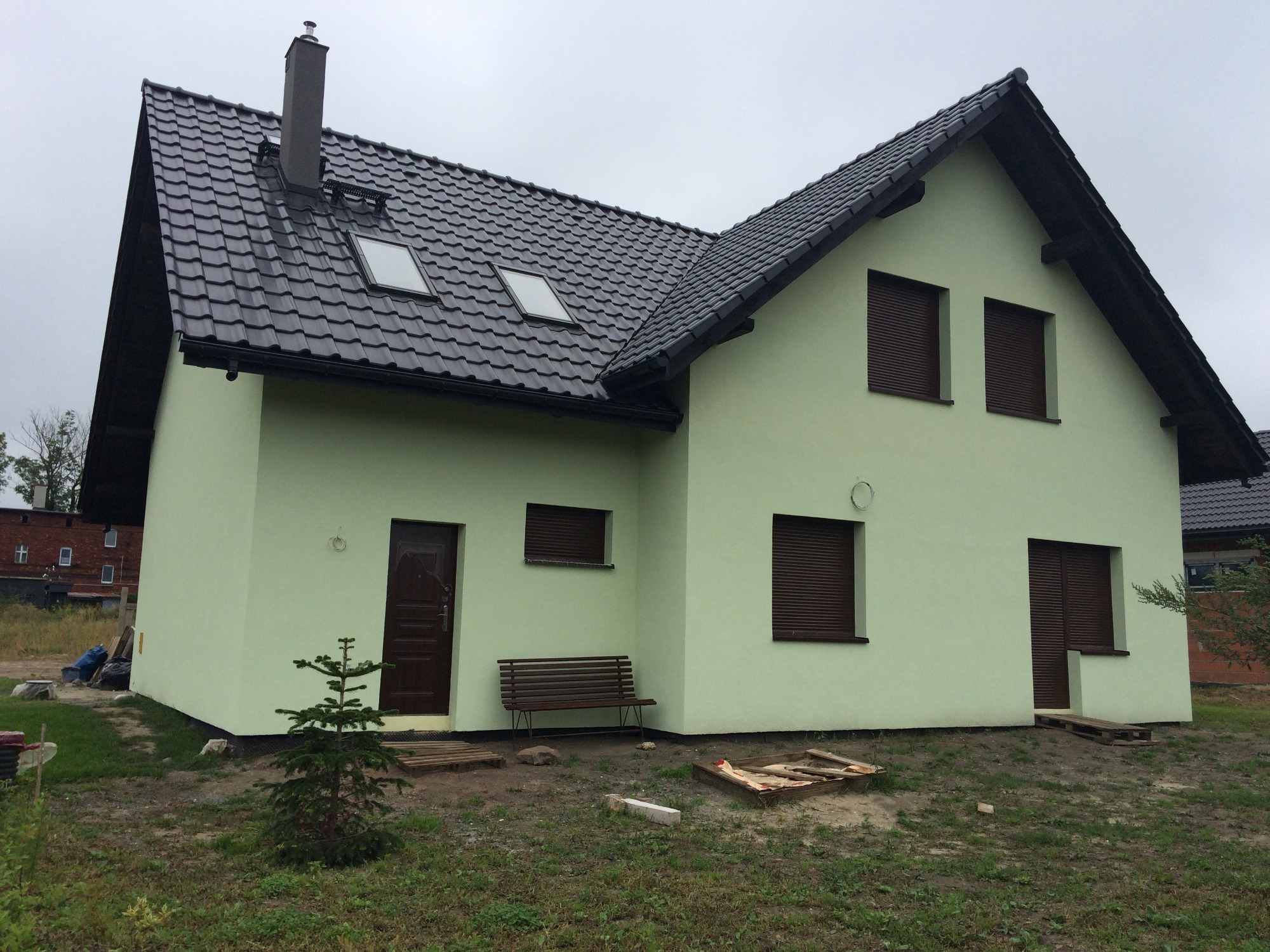 Nowo wybudowany dom murowany z zieloną elewacją, brązowymi roletami i ciemną dachówką, widoczny mały świerk przed wejściem i ławka obok drzwi.