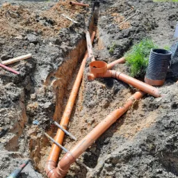 Pomarańczowe rury kanalizacyjne ułożone w wykopie ziemnym, widoczna mini koparka w tle i narzędzie do cięcia rur na pierwszym planie.