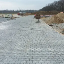 Szeroki, szary plac wyłożony kostką brukową w jodełkę, w tle hałdy ziemi, palety z kostką, maszyny budowlane i drzewa bez liści pod pochmurnym niebem.