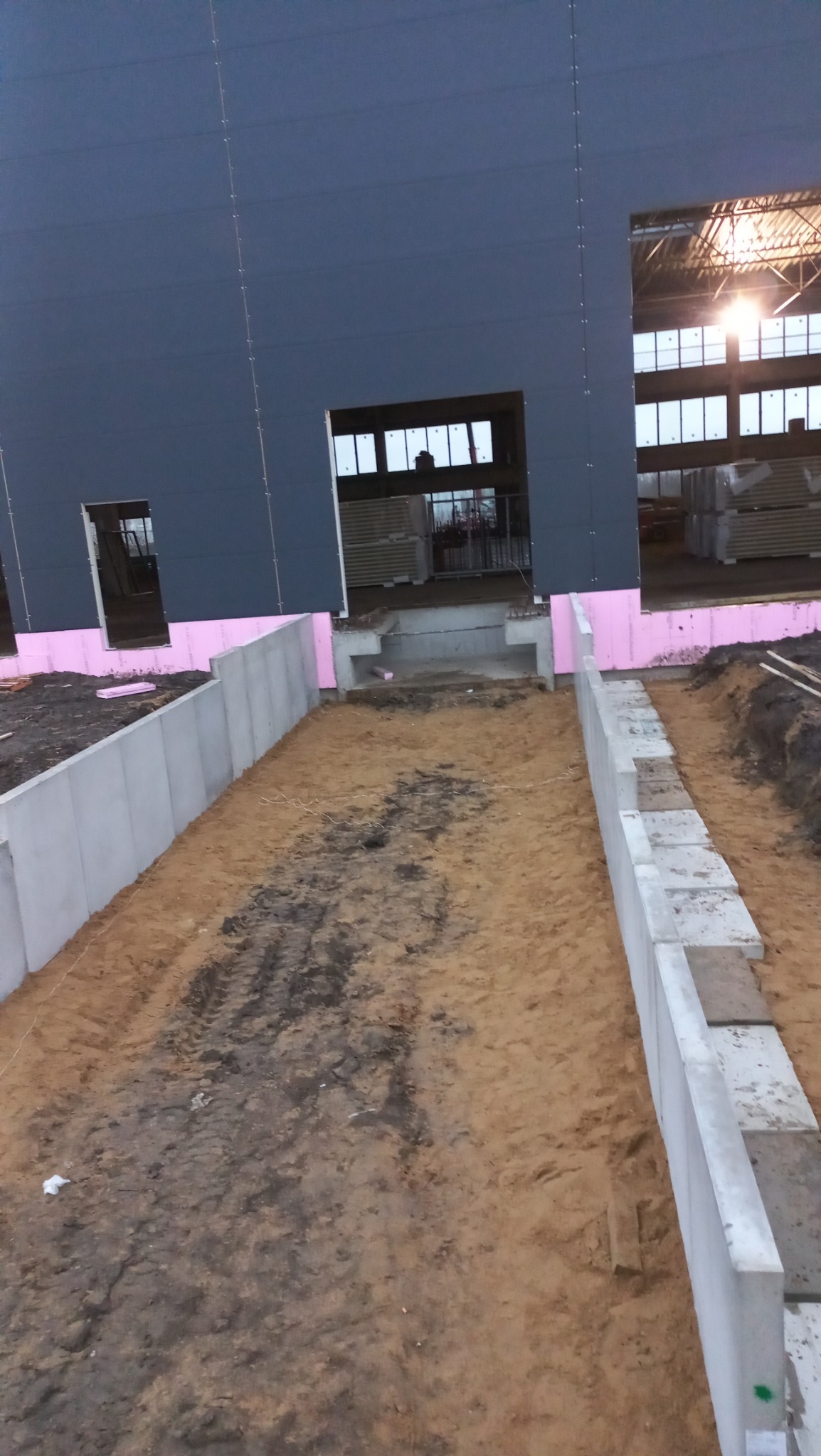 Przygotowany teren pod budowę hali przemysłowej z widocznymi fundamentami i izolacją termiczną, ziemia częściowo utwardzona, w tle widoczne materiały budowlane wewnątrz hali.