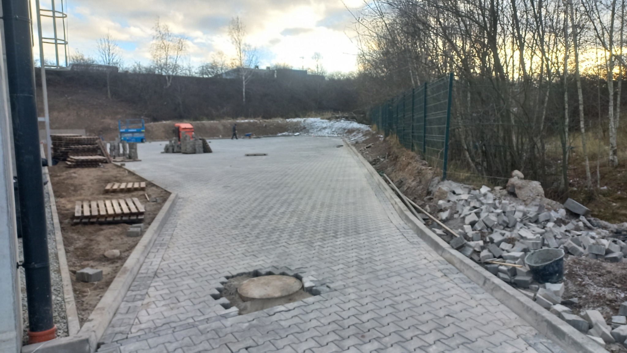 Ułożona szara kostka brukowa na placu z niezakończonym fragmentem wokół studzienki, palety z kostką w tle, teren przygotowany pod dalsze prace.