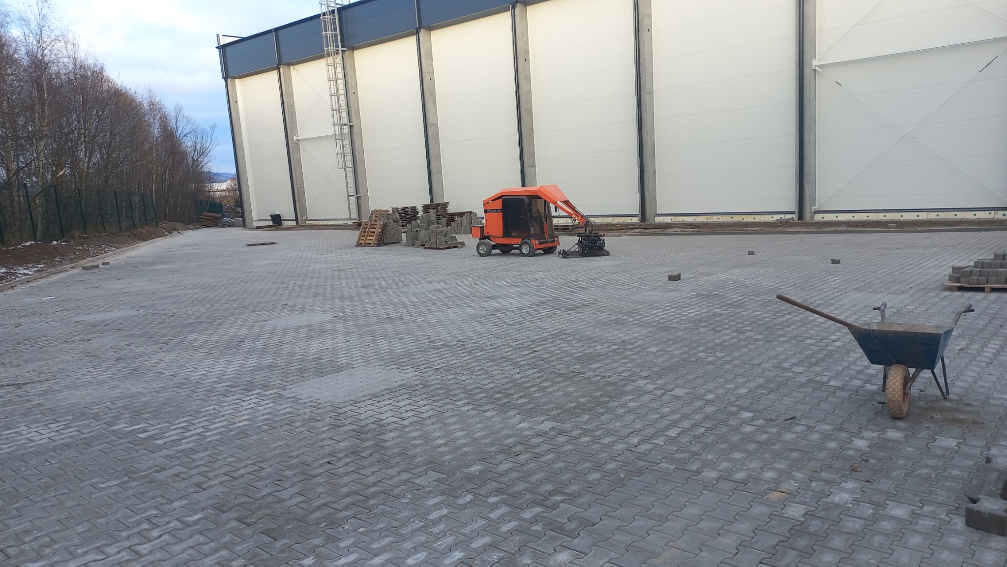 Rozległy plac wyłożony szarą kostką brukową, z widocznym wózkiem budowlanym, pomarańczową maszyną do układania kostki oraz paletami z kostką w tle, na tle hali przemysłowej.