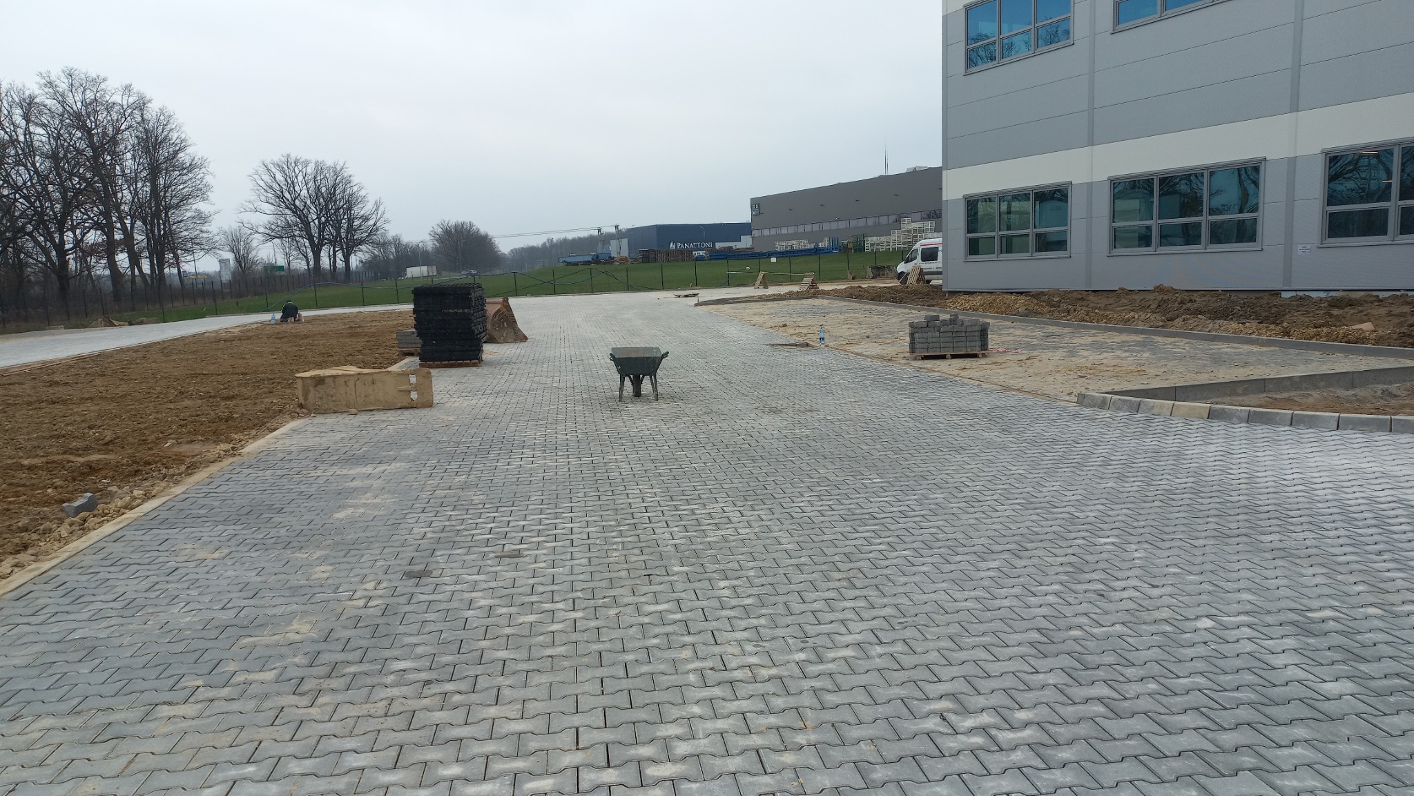 Ukończona nawierzchnia z szarej kostki brukowej typu 'I', widoczny fragment budynku przemysłowego w tle, taczka na środku placu, zapas kostki i pracownik wykonujący prace ziemne z boku.