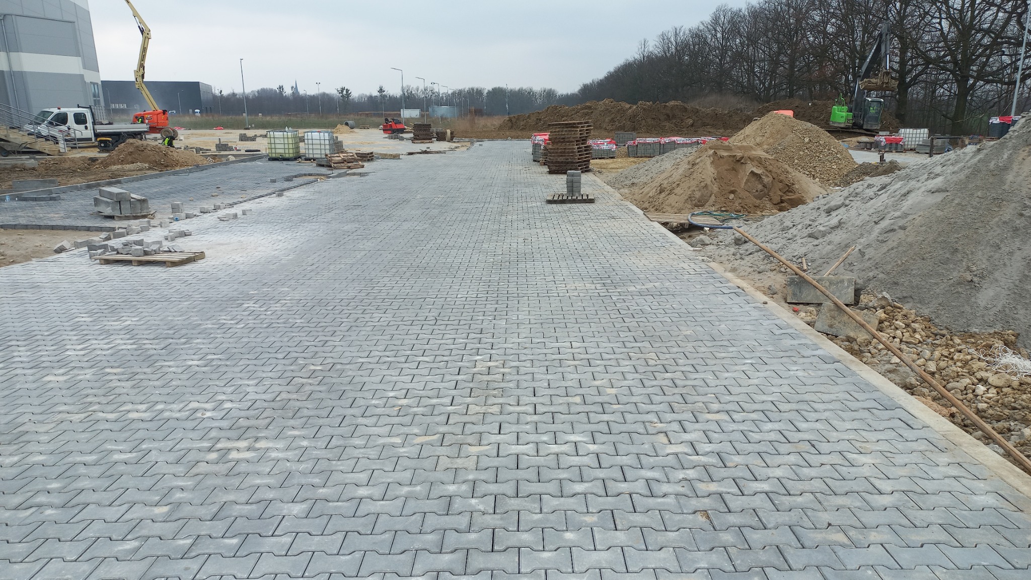 Szeroki, szary plac wyłożony kostką brukową w jodełkę, w tle hałdy ziemi, palety z kostką, maszyny budowlane i drzewa bez liści pod pochmurnym niebem.