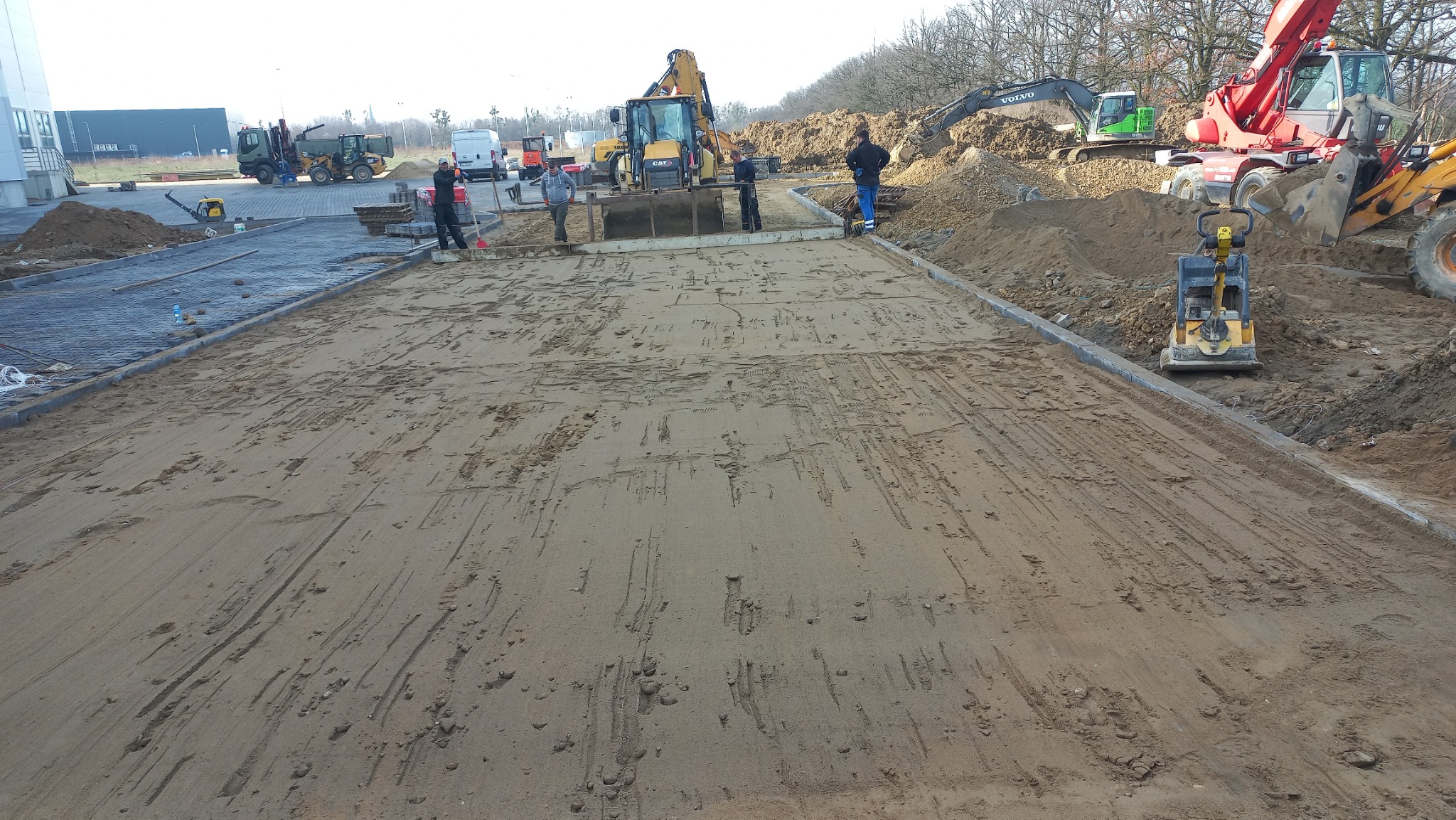 Przygotowanie podłoża pod nawierzchnię: wyrównany teren z obrzeżami, ubijarka wibracyjna, ekipa robotników i ciężki sprzęt budowlany w tle.