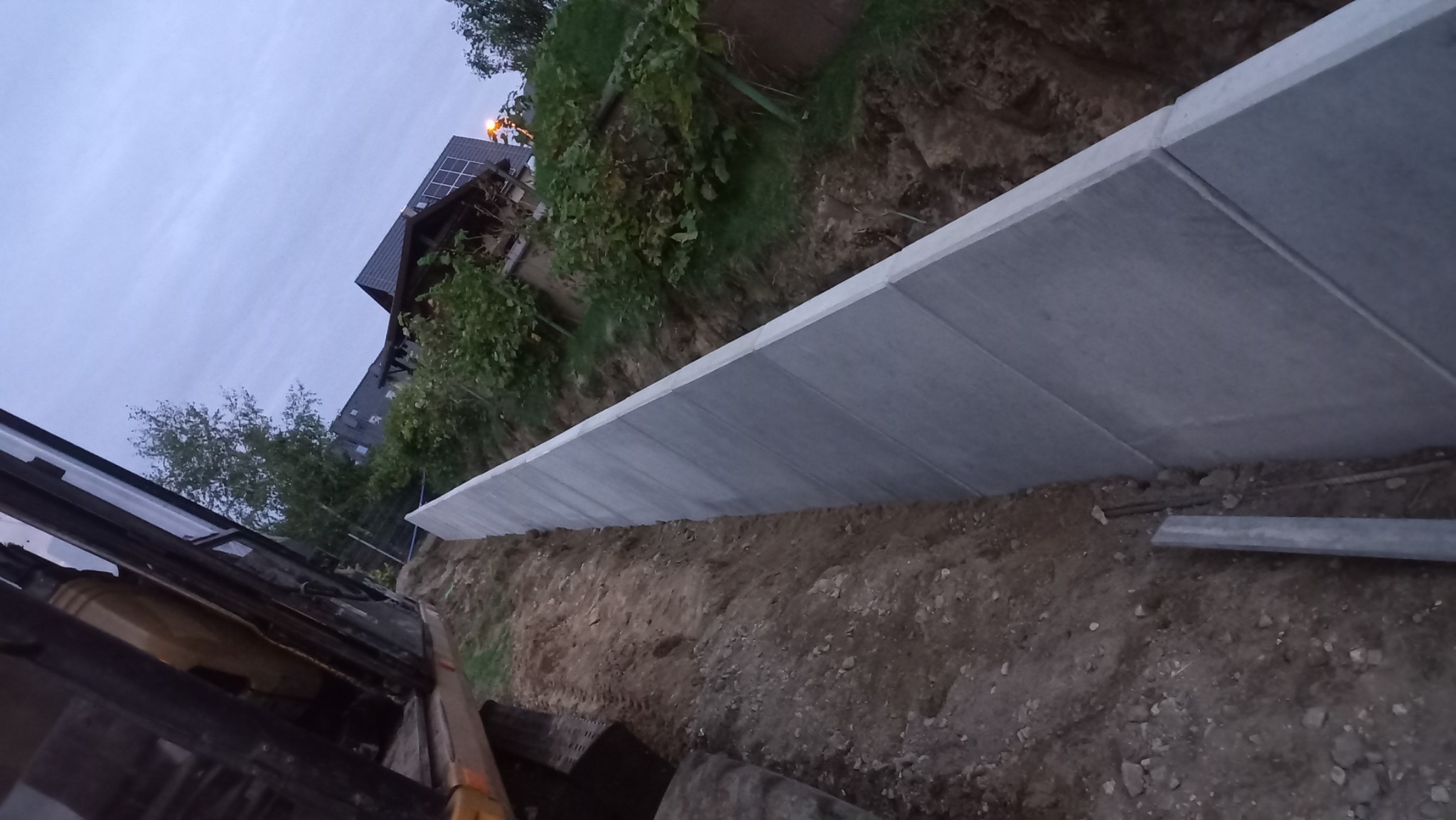 Wzmocnienie skarpy betonowymi płytami, widok z kabiny maszyny budowlanej, w tle dom z panelami słonecznymi.