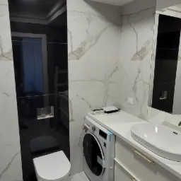 Nowoczesna łazienka z biało-czarnymi płytkami imitującymi marmur, wisząca toaleta, pralka pod blatem i umywalką z szafką z szufladami w kolorze białym.