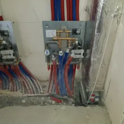 Rozdzielacz instalacji grzewczej z podłączonymi rurami w kolorze czerwonym i niebieskim, zamontowany na ścianie w surowym stanie, widoczne liczniki i izolacja termiczna kanału wentylacyjnego...