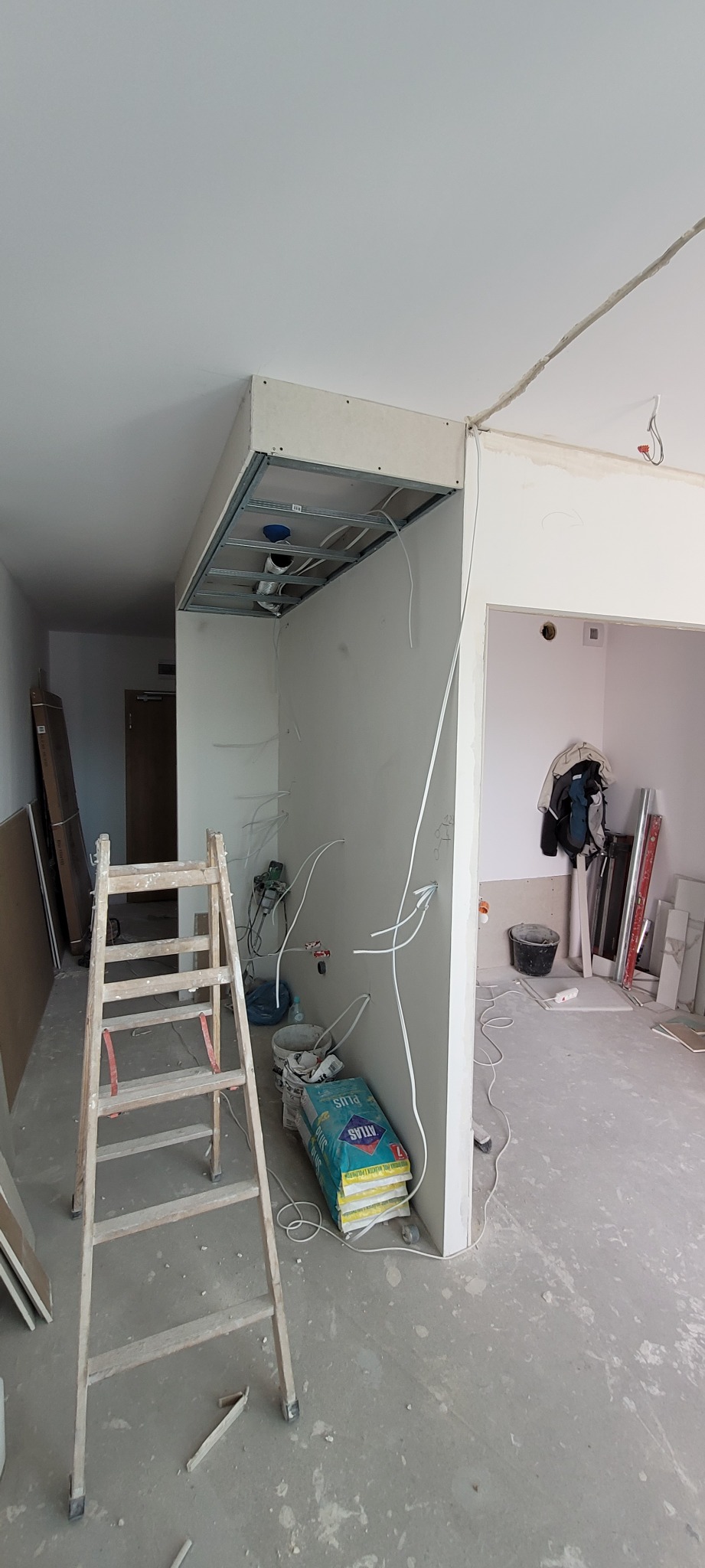 Wnętrze w trakcie remontu: widoczna konstrukcja sufitu podwieszanego z instalacjami, ściany z płyt gipsowo-kartonowych, drabina, worki z gładzią Atlas Plus oraz kable elektryczne wystające ze ścian.
